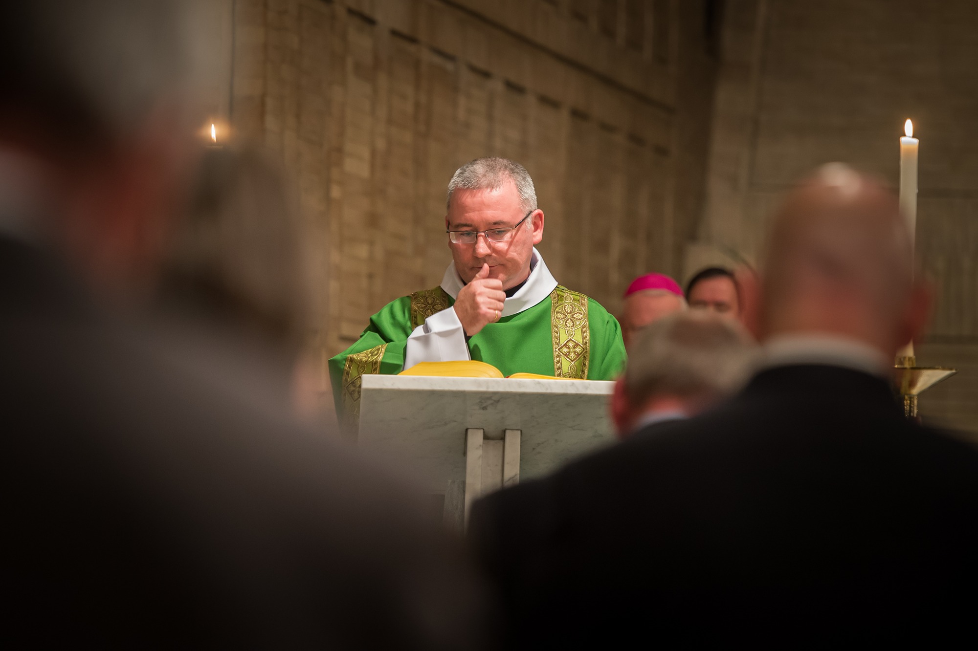 Priest in green vestments standing at a marble lectern with an open book, engaged in a religious service inside a church.