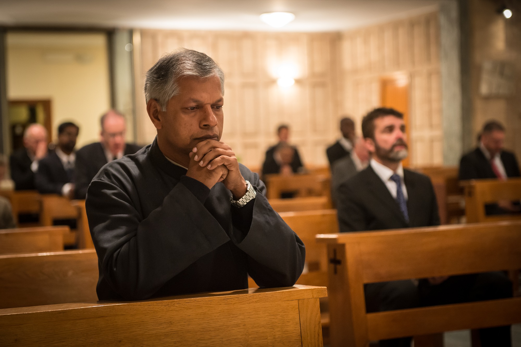 Man in clerical attire praying with folded hands in a church pew, surrounded by other seated people.