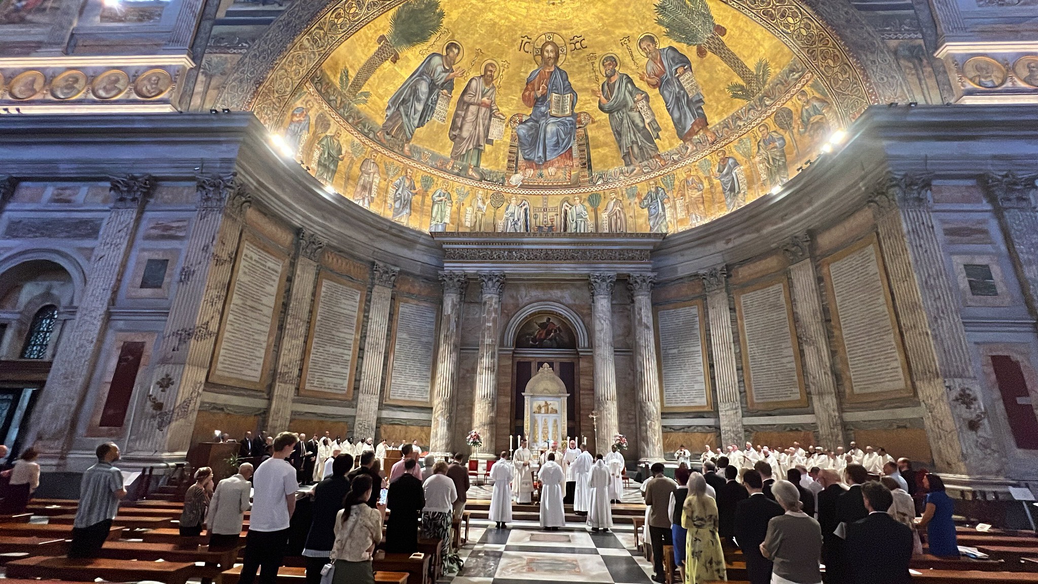 Interior of a large church with people attending a religious ceremony, featuring a golden mosaic dome depicting Jesus and saints.