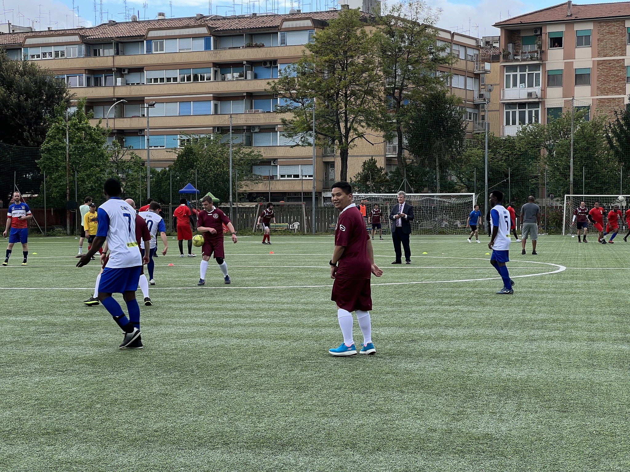Group of people playing soccer on a green artificial turf field in an urban area with apartment buildings in the background.