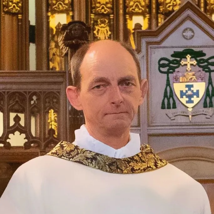 Middle-aged clergyman wearing white and gold ornate vestments standing inside a church with wooden and stained glass background.