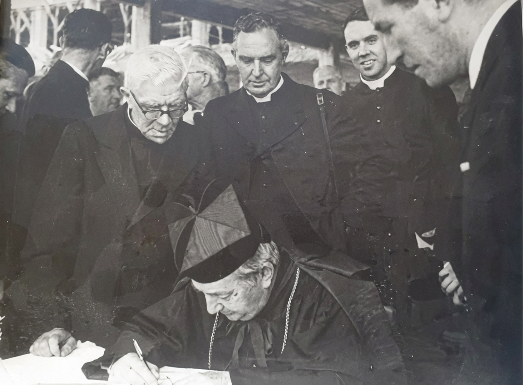 Black and white photo of a clergyman in ceremonial attire signing a document surrounded by other clergymen.