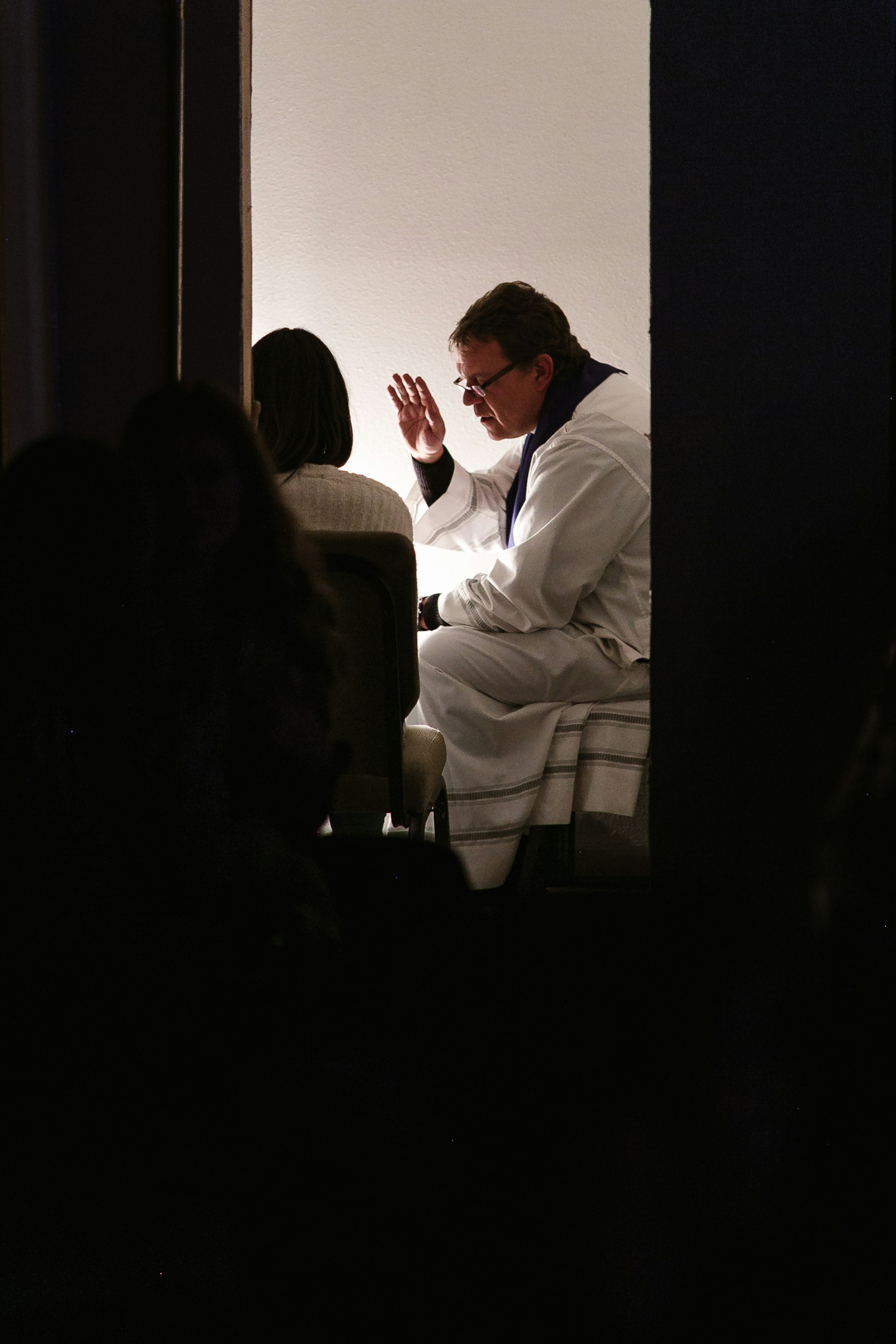 A priest in white robes with a purple stole is raising his hand to bless or speak to a seated woman in a dimly lit room.