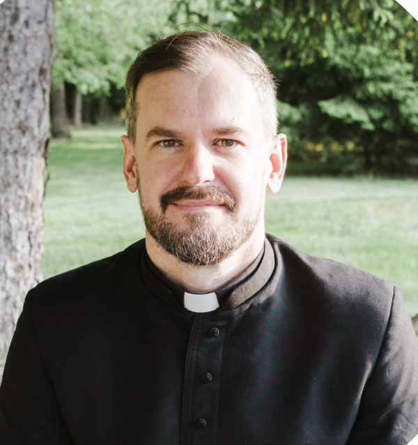 Portrait of a man with a beard wearing a black clergy shirt with a white clerical collar standing outdoors near a tree.
