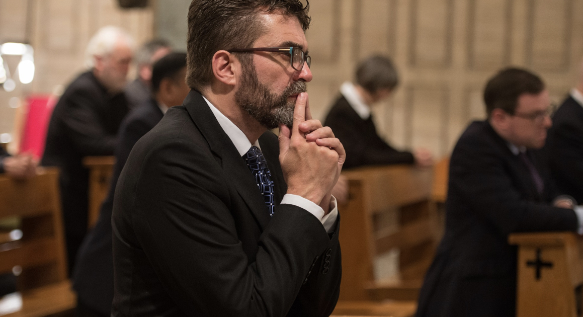 Man in glasses and suit sitting in a church pew with hands clasped near his mouth, appearing thoughtful or prayerful.