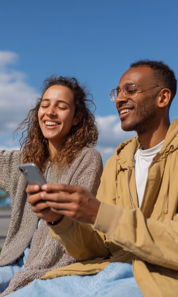 Smiling young woman and man sitting outdoors, looking at a smartphone together under blue sky.