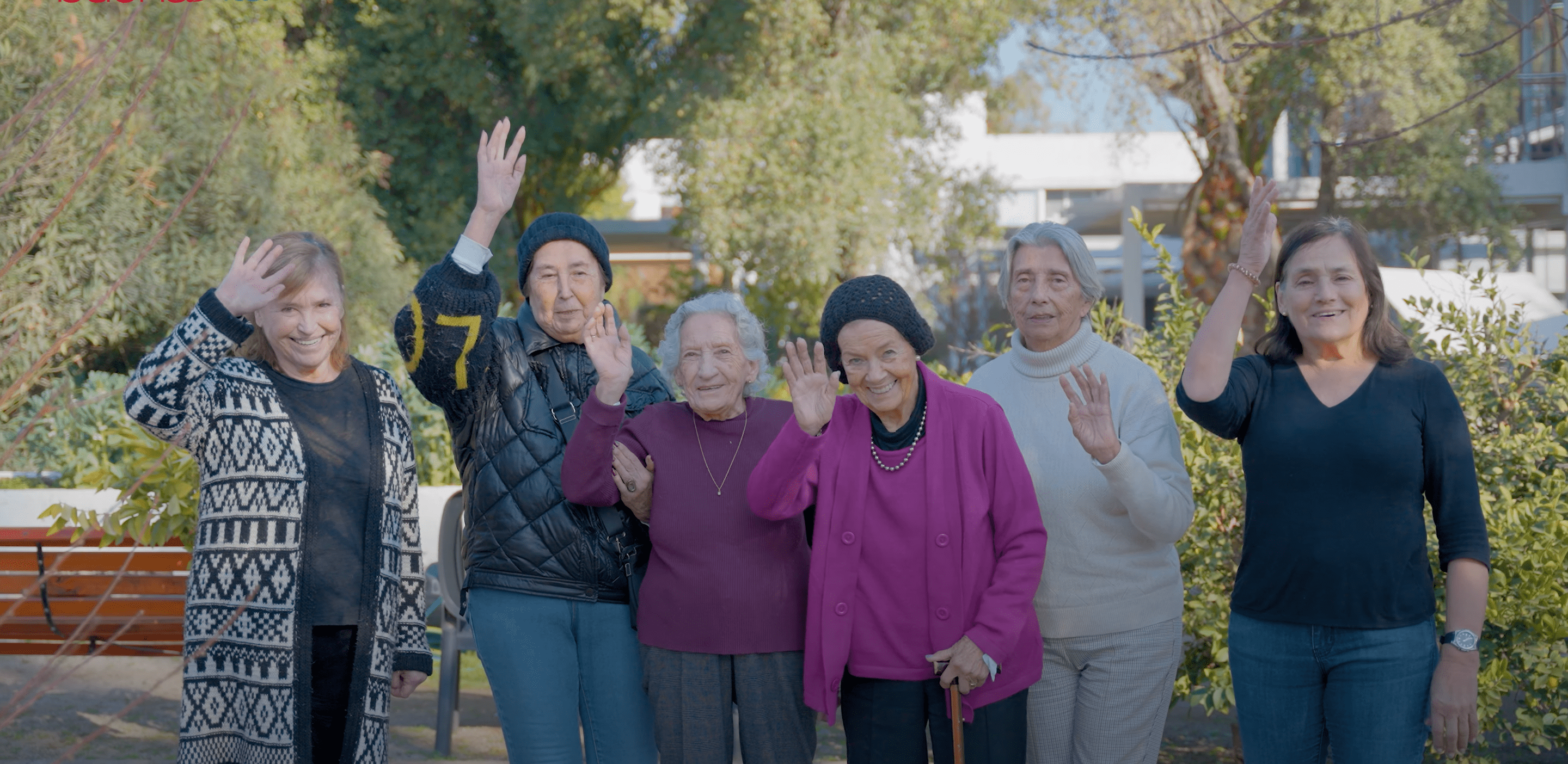 Seis mujeres mayores sonrientes saludando al aire libre en un parque con árboles y arbustos alrededor.