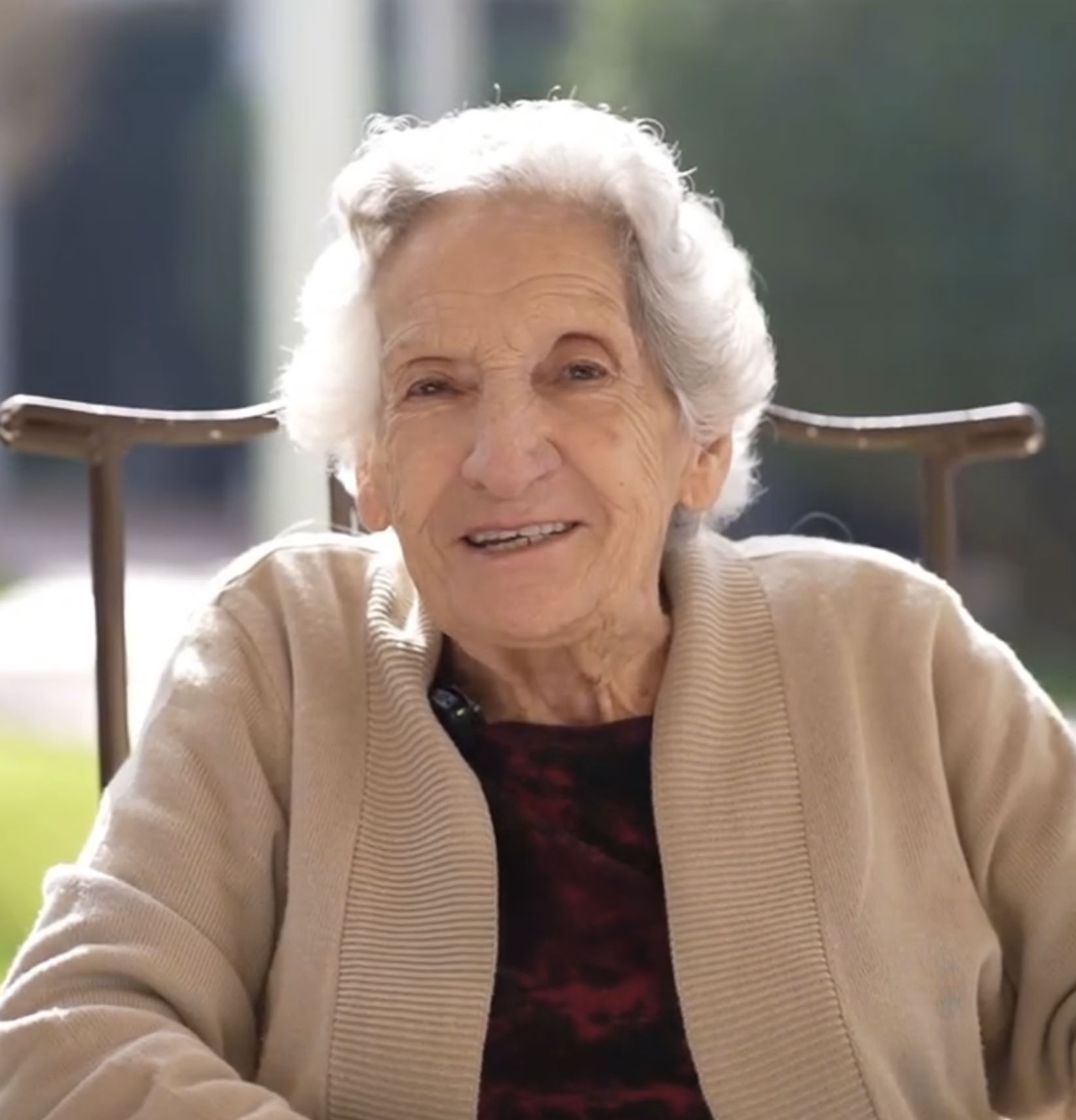 Mujer mayor con cabello canoso y suéter beige sonriendo mientras está sentada al aire libre.