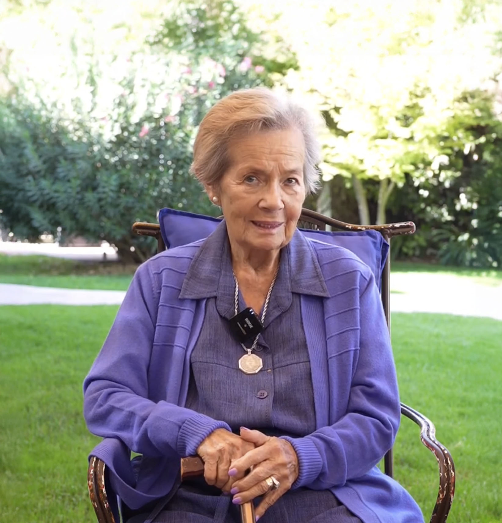 Mujer mayor sentada en una silla con suéter y blusa morados, sosteniendo un bastón en un jardín con césped y árboles.