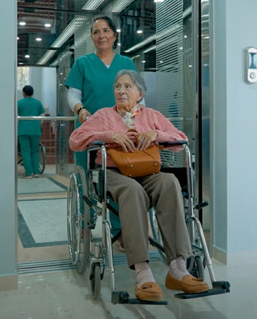 Trabajadora de salud empujando a una mujer mayor en silla de ruedas dentro de un edificio moderno.