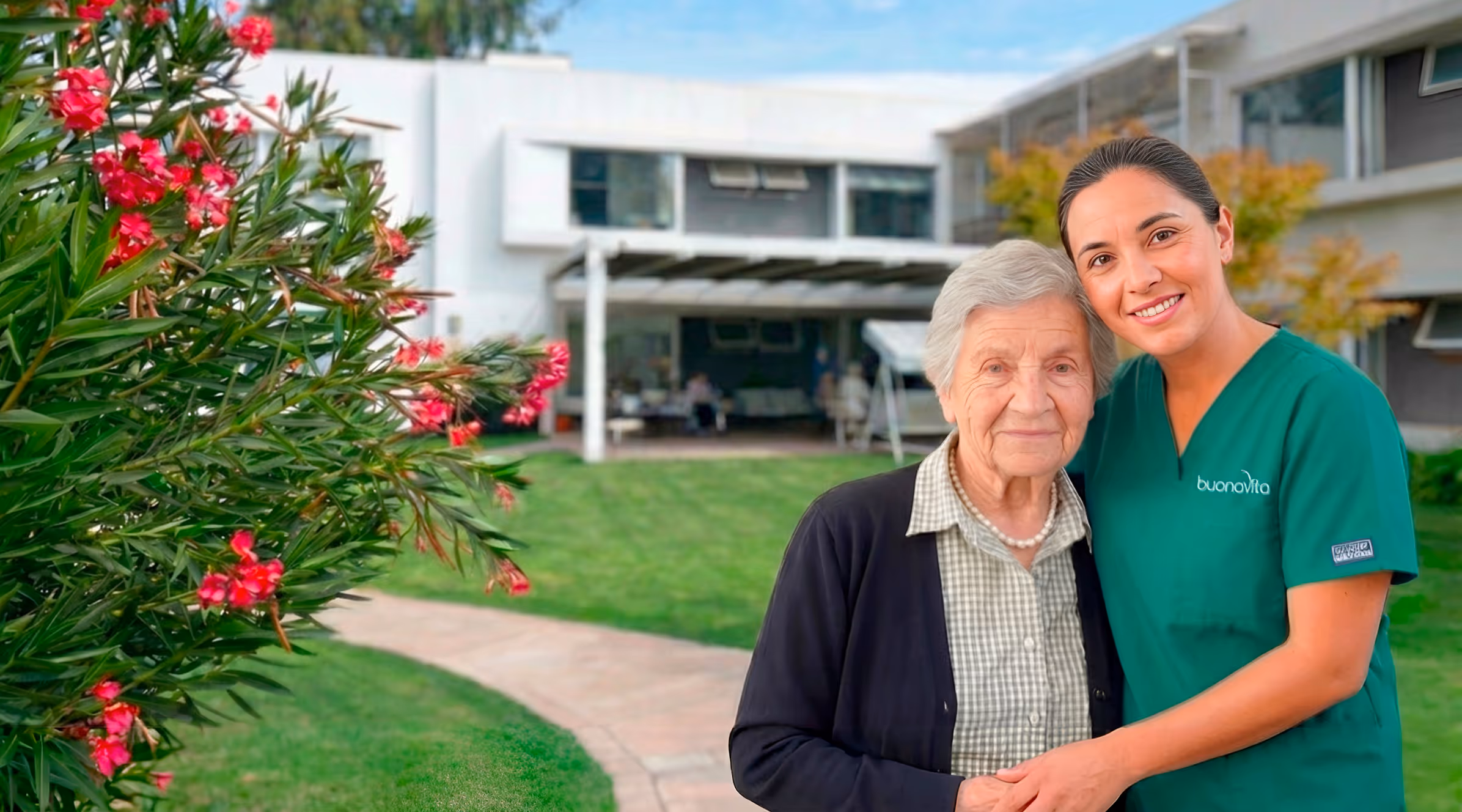 Mujer mayor con su cuidadora sonriente abrazándose en un jardín frente a un edificio moderno.