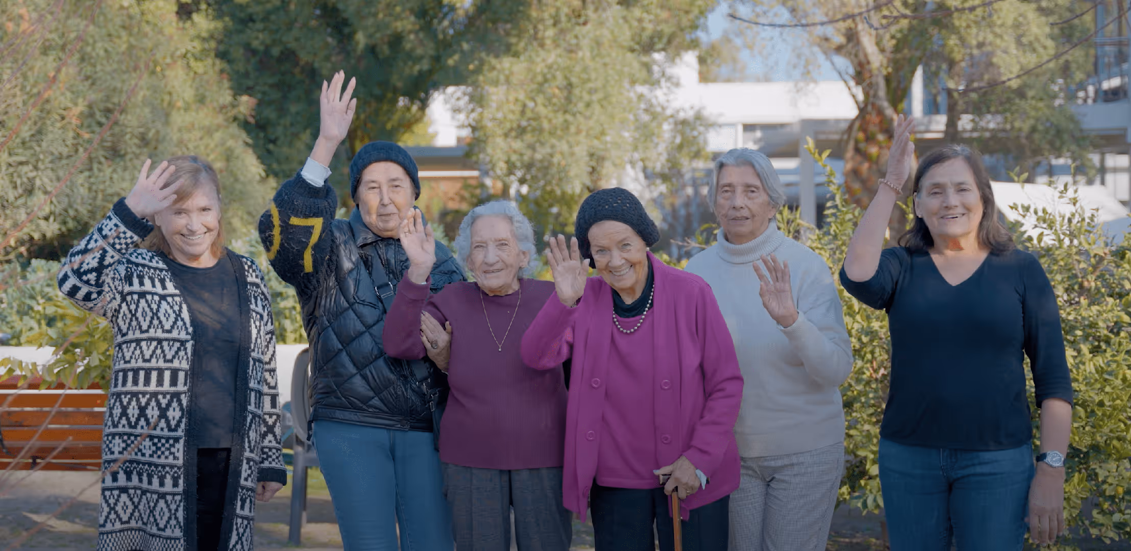 Seis mujeres mayores sonrientes saludando al aire libre en un parque con árboles y arbustos alrededor.