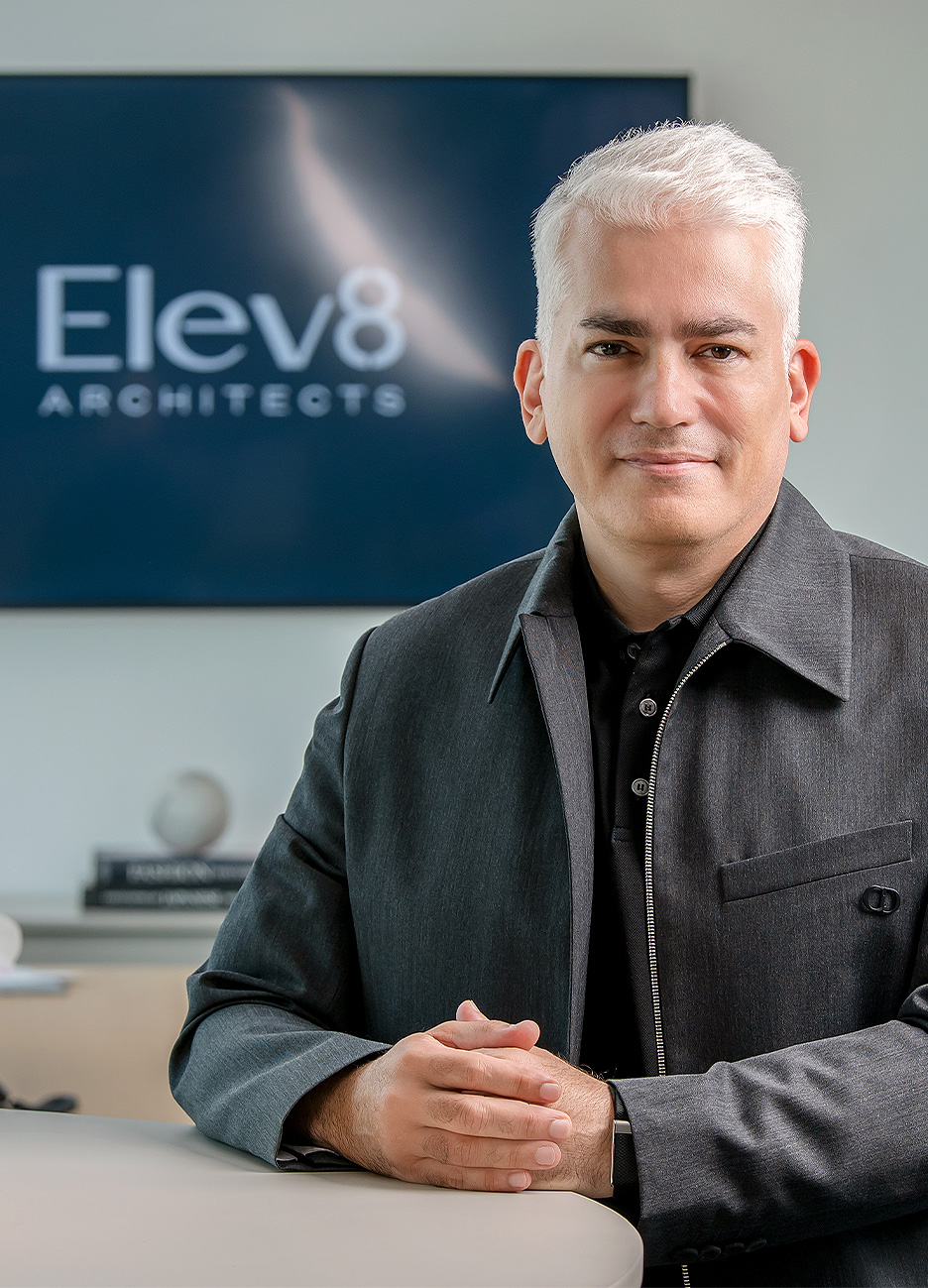 Man with short white hair wearing a dark jacket sitting at a table with hands folded, with a blurred screen in the background displaying 'Elev8 Architects.'