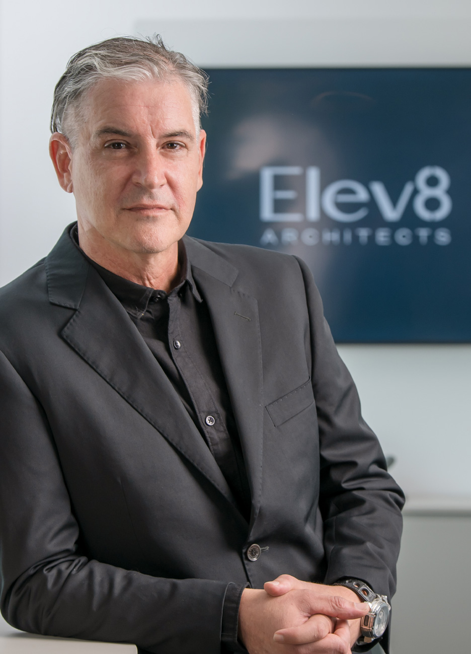 Middle-aged man in a black blazer and shirt, seated with hands clasped, in front of a blurred screen displaying 'Elev8 Architects'.