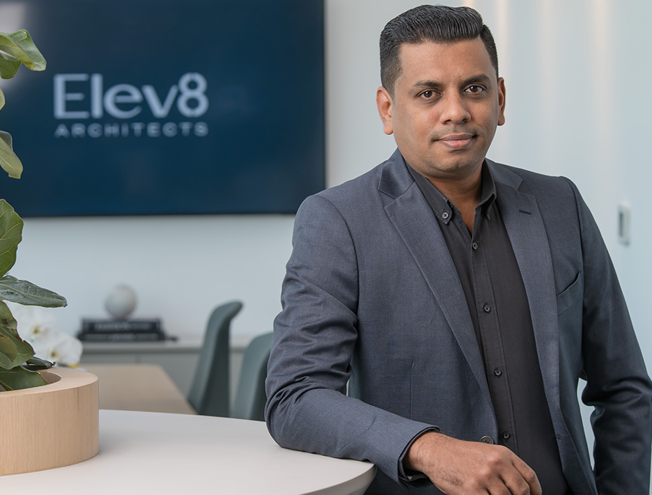 Man in a dark blazer and black shirt leaning on a white desk with a blurred background featuring a sign that reads 'Elev8 Architects'.