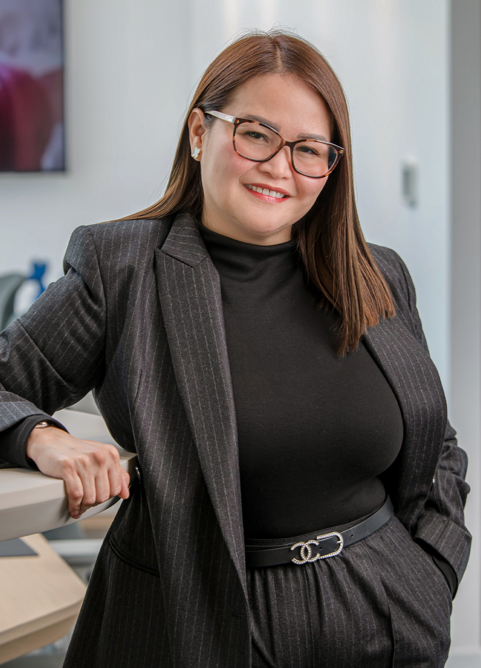 Smiling woman with shoulder-length brown hair, wearing glasses, a dark pinstripe blazer, black top, and black belt with a decorative buckle, leaning on a desk in a modern office.