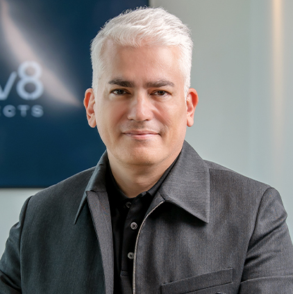 Man with white hair wearing a dark jacket, sitting indoors with a blurred logo in the background.
