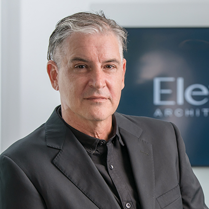 Portrait of a middle-aged man with gray hair wearing a black suit jacket and shirt, standing indoors with a blurred blue sign in the background.