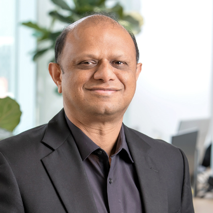 Man wearing a black suit jacket and shirt smiling in an office setting with blurred plants and computers in the background.