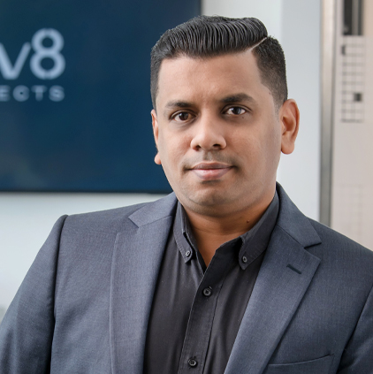 Professional man wearing a dark suit jacket and black shirt, standing in an office with blurred background showing part of a logo.