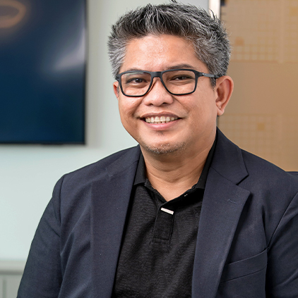 Smiling man with gray hair and black glasses wearing a dark blazer and black shirt in an indoor office setting.