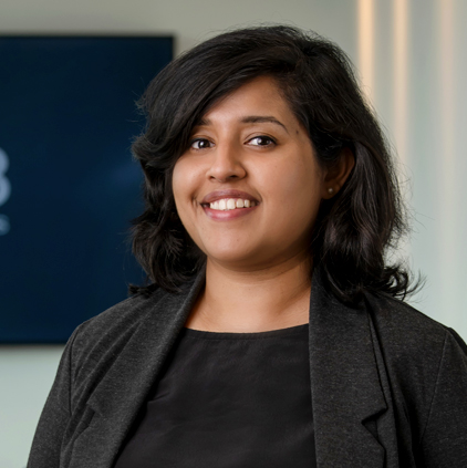 Portrait of a smiling woman with shoulder-length black hair wearing a black top and dark gray blazer, standing indoors.