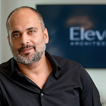 Man with short beard and receding hairline wearing a black shirt, posing indoors with a blurred sign reading 'Elev' in the background.