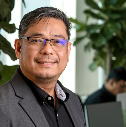 Smiling man wearing glasses and a dark suit jacket in an indoor office setting with plants and a blurred person in the background.