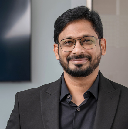 Man with beard and glasses wearing a black suit jacket and shirt smiling against a blurred indoor background.