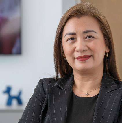 Confident woman with long brown hair wearing a black pinstripe blazer and gold jewelry, smiling in an office setting.