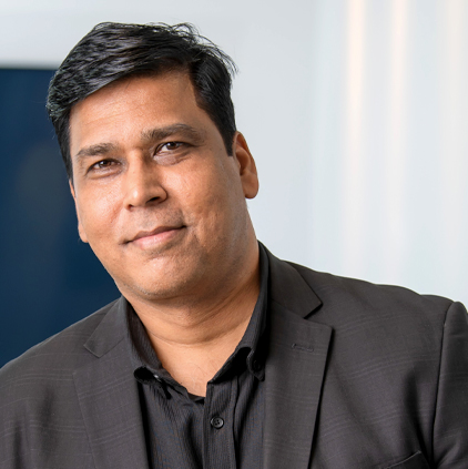 Middle-aged man with dark hair wearing a black suit jacket and black shirt, smiling slightly in an indoor setting.