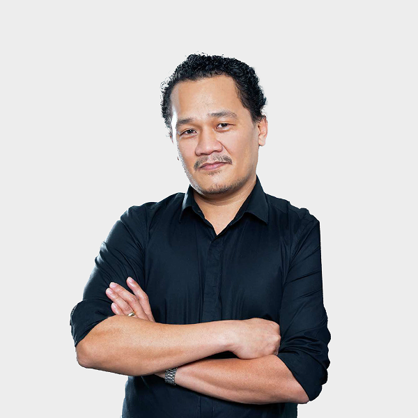 Man with curly hair and mustache wearing a black shirt, standing with arms crossed against a plain light background.