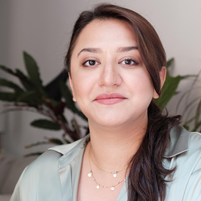 Portrait of a woman with dark hair tied back, wearing a light blue shirt and layered gold necklaces, against a background with green plants.