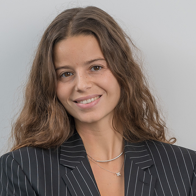 Smiling young woman with long wavy brown hair wearing a black pinstripe blazer and layered necklaces, posing against a plain light background.