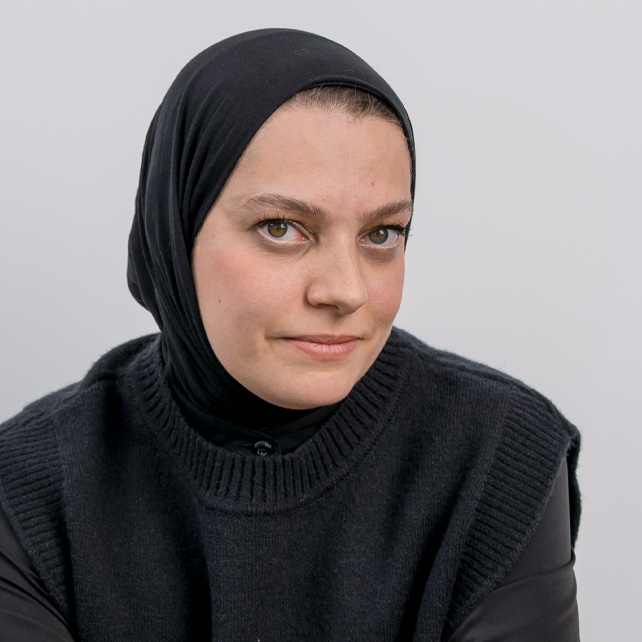 Close-up portrait of a woman wearing a black hijab and a dark knitted sleeveless sweater looking at the camera.