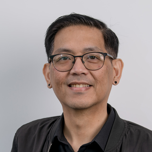 Smiling man with short black hair, wearing glasses, black earrings, and a dark jacket over a black shirt against a plain light gray background.