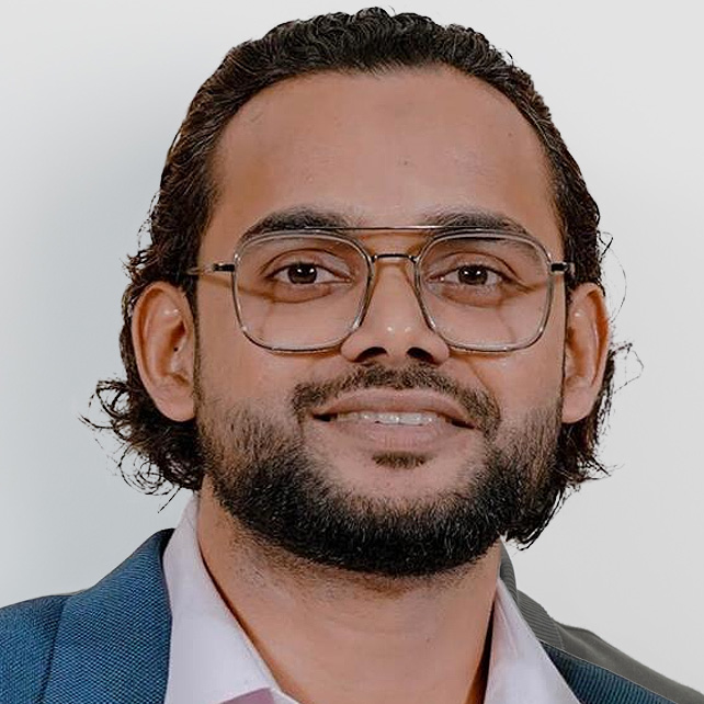 Smiling man with glasses, beard, and curly hair wearing a blue suit jacket and light pink shirt.