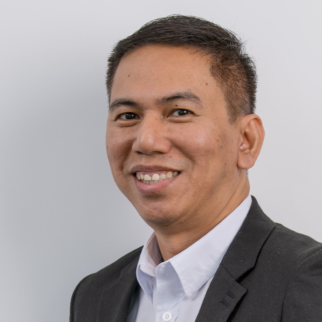 Smiling man in a dark blazer and white collared shirt against a plain light gray background.