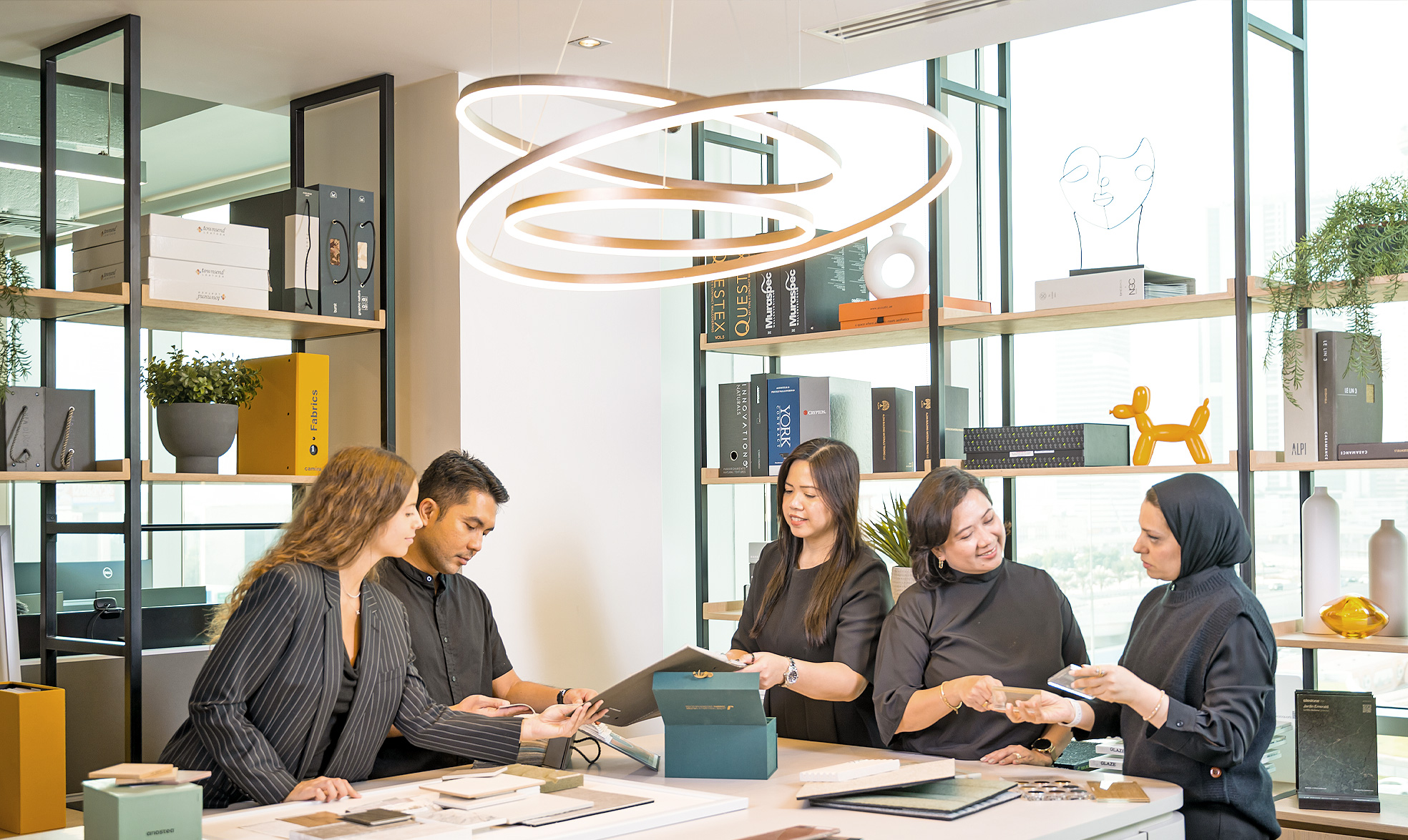 Five professionals in a modern office reviewing materials and samples at a table under circular pendant lights.
