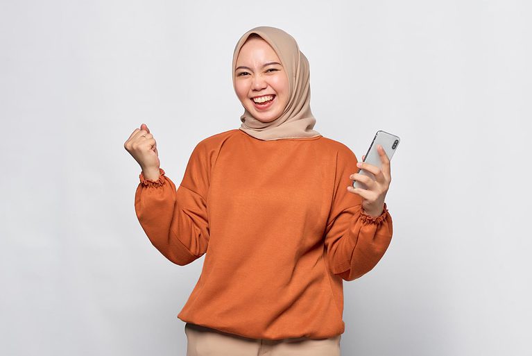 Smiling woman wearing a beige hijab and orange sweater holding a smartphone and making a fist-pump gesture.