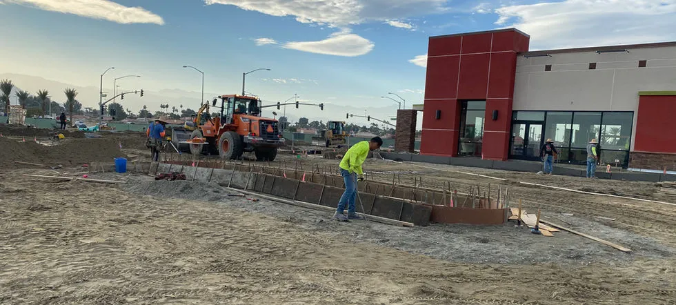 Construction site with workers and machinery preparing the ground in front of a new commercial building with a red and beige facade.