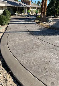Curved stamped concrete driveway leading to a residential house with trimmed bushes and palm trees.