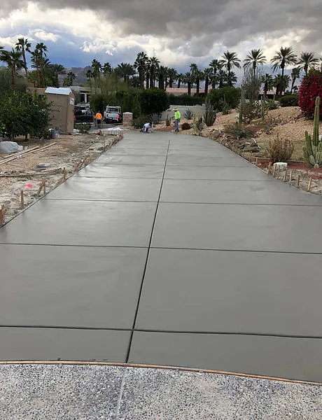 Freshly poured and smoothed concrete driveway under construction with workers in the background and palm trees lining the sides.