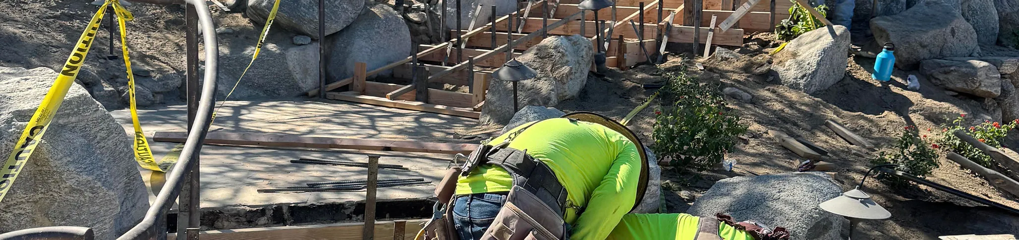 Construction worker in yellow shirt and hard hat working on concrete formwork surrounded by rocks and caution tape.