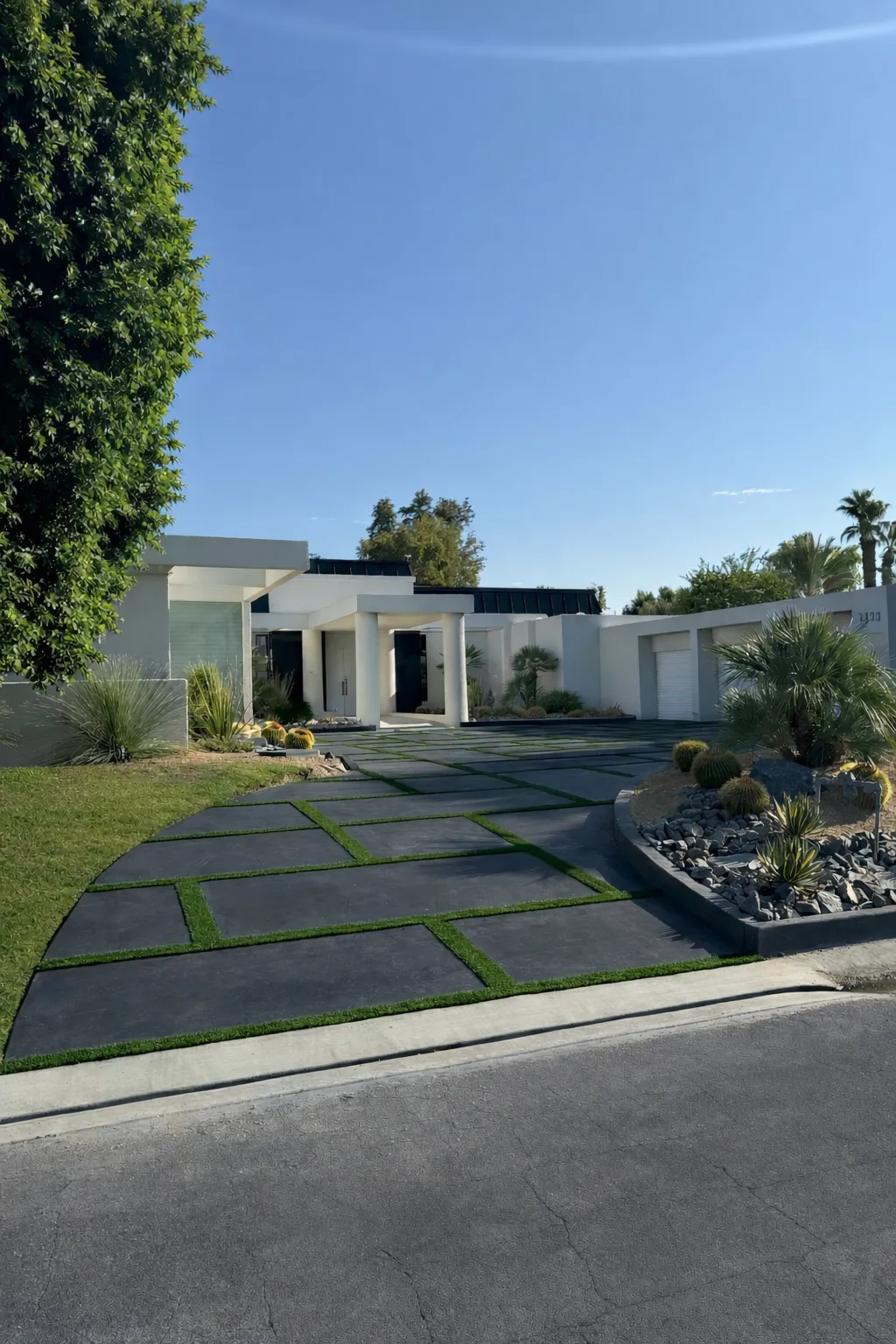 Modern white house with a driveway featuring dark paving stones separated by green grass strips and desert landscaping with cacti and rocks.