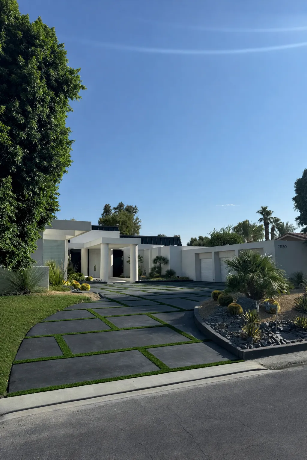 Modern white house with a driveway made of large rectangular concrete slabs separated by strips of green grass, surrounded by desert landscaping and trees under a clear blue sky.