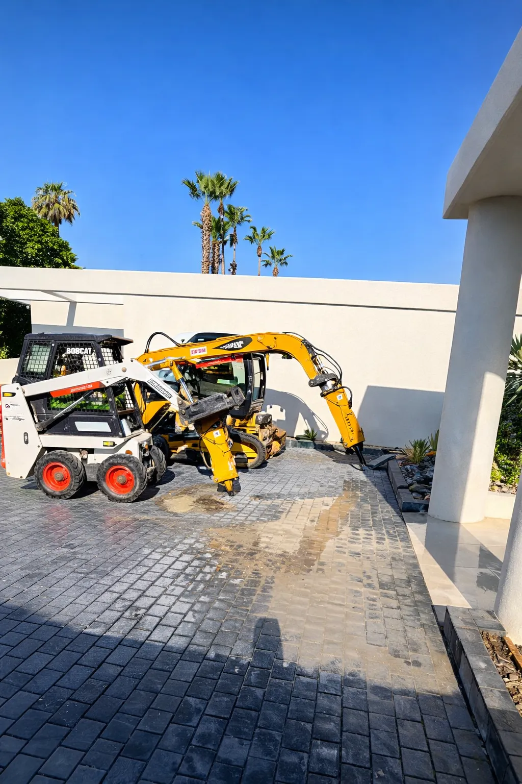 Small Bobcat skid-steer loader and yellow excavator parked on a paved driveway near a white building with palm trees in the background.