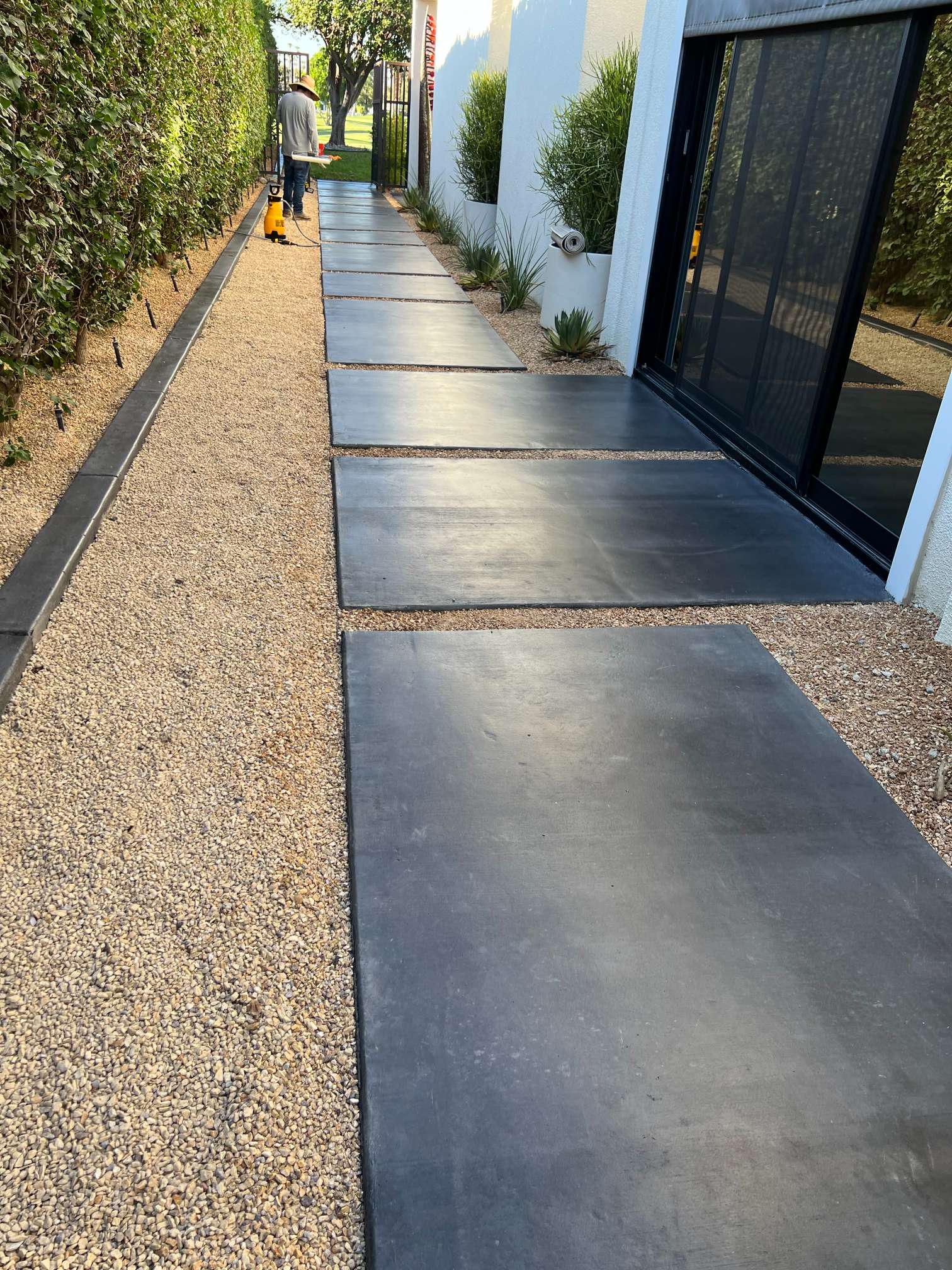 Person working on a narrow garden pathway with large rectangular concrete stepping stones and gravel surrounded by hedges and a white building.
