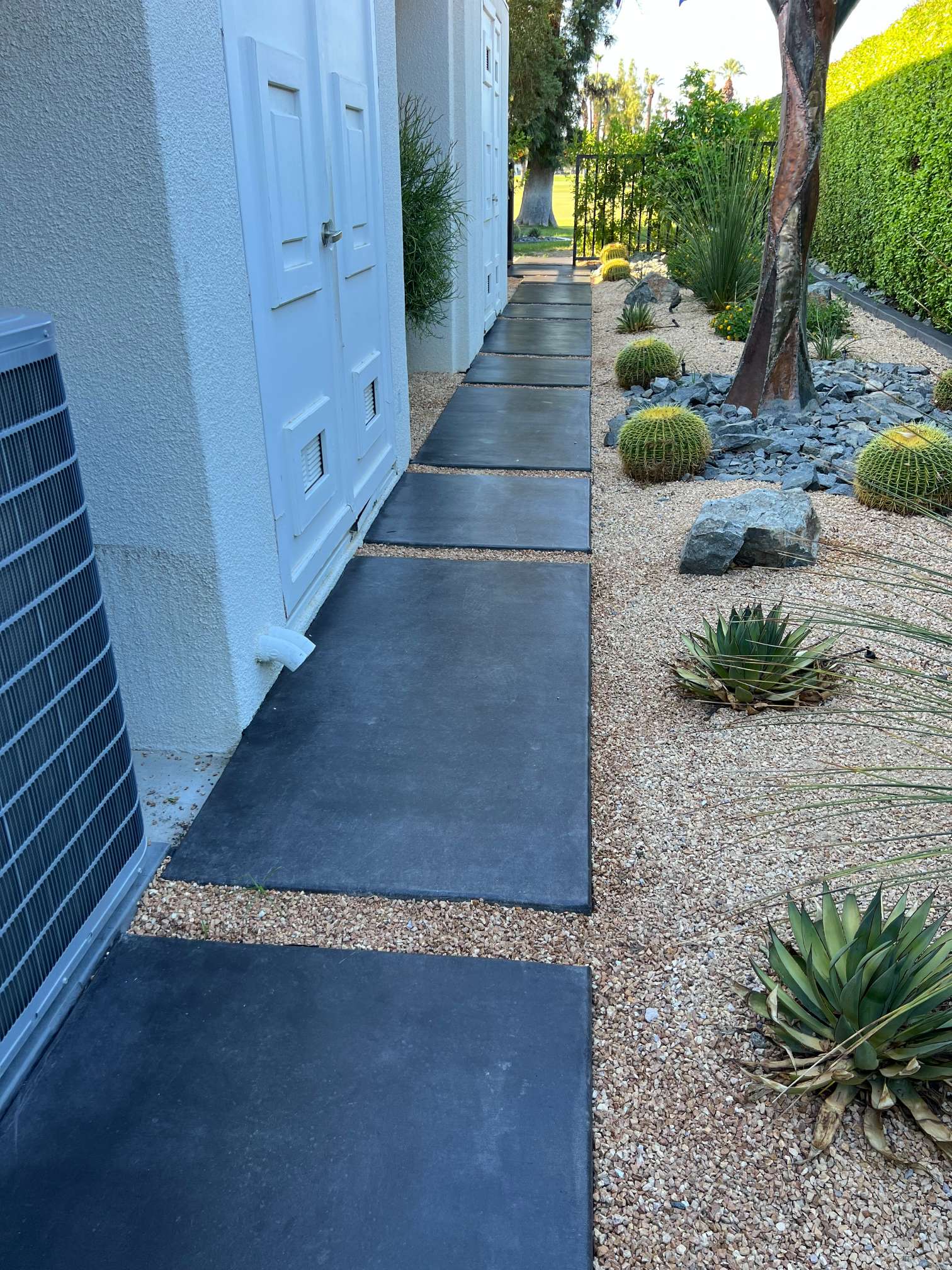 Pathway with large black square stepping stones alongside a white building, bordered by a desert garden with cacti, gravel, and a trimmed green hedge.