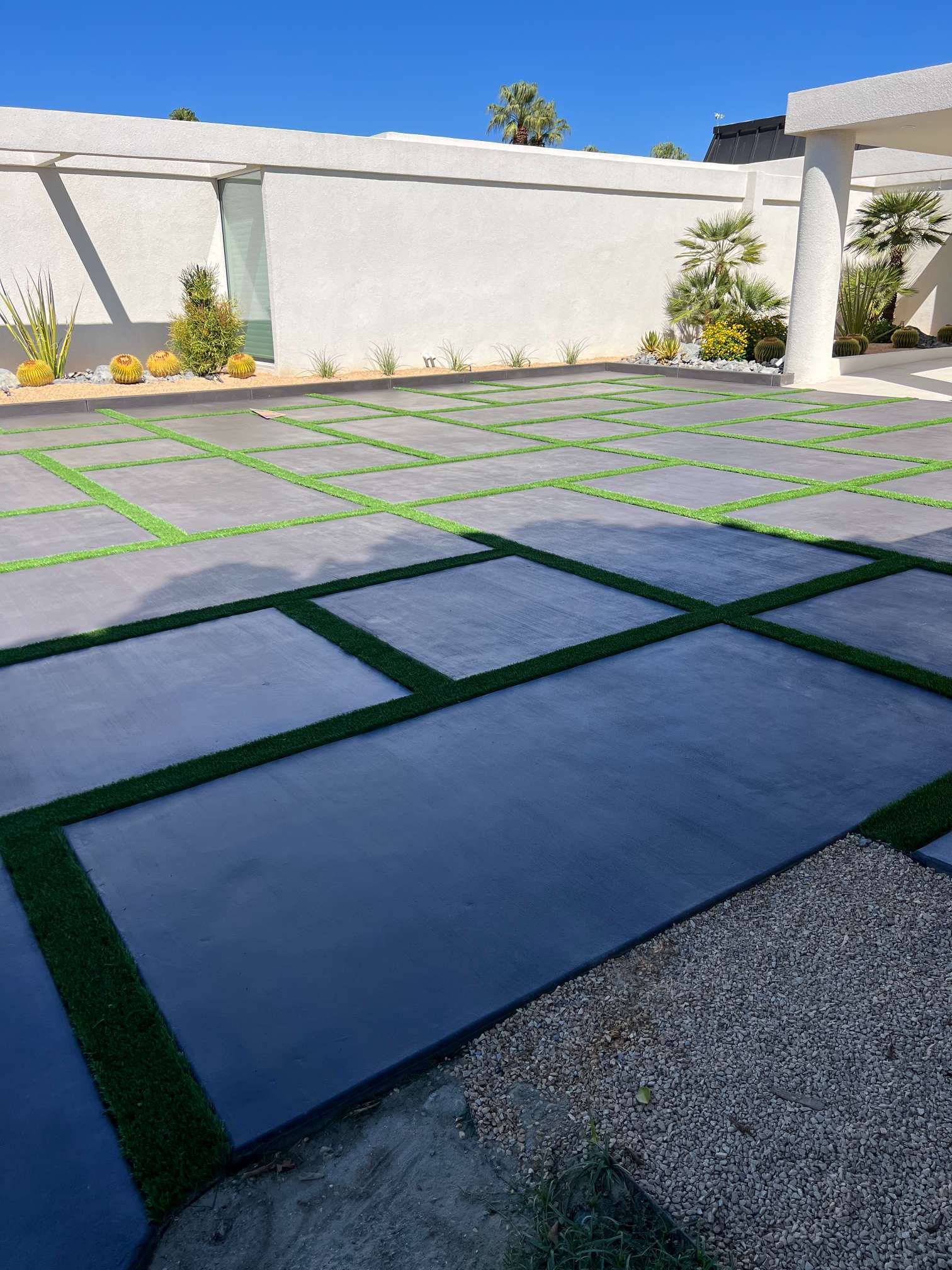 Modern outdoor patio with large square concrete slabs separated by strips of green grass, surrounded by desert plants and a white wall under a clear blue sky.